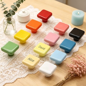 May include: A collection of colorful square cabinet knobs in various shades, including red, orange, yellow, green, pink, black, blue, and white. Some knobs have names printed on them. The knobs are arranged on a wooden surface.