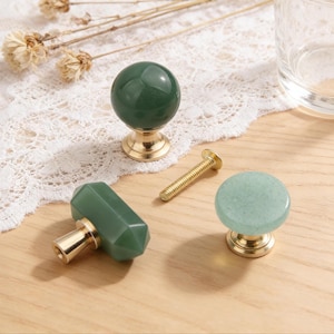 Puede incluir: Un conjunto de pomos de armario de piedra verde con bases de latón y un tornillo de latón. Los pomos tienen diferentes formas: una esfera, un cilindro y un círculo plano. Los pomos están sobre una superficie de madera clara.