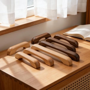 Può includere: Una collezione di maniglie per mobili in legno in varie tonalità di marrone, dal chiaro allo scuro. Le maniglie sono arcuate e hanno una finitura liscia e lucida. Sono disposte su una superficie di legno, mostrando diversi stili.