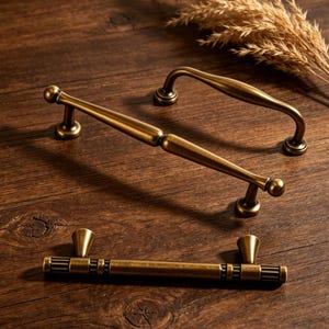 May include: Three antique brass cabinet pulls and handles are arranged on a wooden surface. The handles have a vintage aesthetic, with a curved bar handle and two different styles of pulls. The hardware is a warm, aged brass colour.
