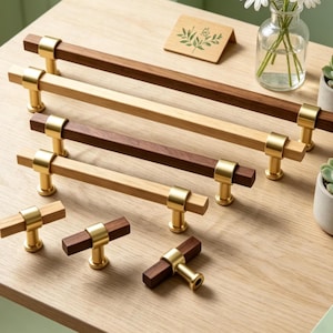 May include: A collection of wooden cabinet pulls and knobs with brass accents. The pulls are rectangular with a dark brown or light wood finish. The knobs have a T-shape design. The hardware is displayed on a light wood surface.
