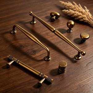 May include: An assortment of antique brass cabinet hardware, including handles and knobs. The handles have curved and straight designs, while the knobs are round and cylindrical. The hardware is arranged on a dark wood surface, with a dried plant in the background.