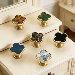 May include: Six decorative cabinet knobs with a cloverleaf design. The knobs feature gold-colored bases and tops in various colors, including amber, green, black, navy blue, and a speckled pattern. The knobs are arranged on a cream-colored surface.