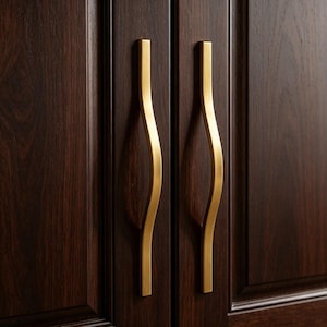 May include: Close-up of two gold-colored cabinet handles with a curved, wave-like design. The handles are mounted on a dark brown wooden cabinet door. The handles have a rectangular base and a smooth, polished finish.