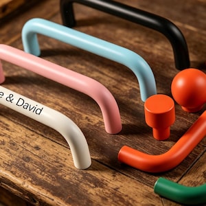 May include: A collection of colorful cabinet hardware, including curved handles in black, light blue, pink, and white, with the text "Alice & David" on one. Also shown are orange knobs and handles, and a green handle. The hardware is displayed on a wooden surface.