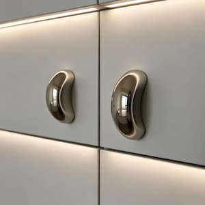 May include: Close-up of two polished, silver-toned cabinet pulls. The pulls have a smooth, curved design and are mounted on a white cabinet door. The pulls reflect the surrounding environment, and are positioned above a soft, warm light.