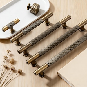 May include: A collection of antique brass cabinet hardware, including four long bar pulls, a small T-shaped pull, and a round knob. The pulls have a ribbed center section and are arranged on a light wood surface.