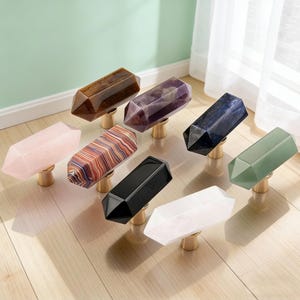 May include: A collection of crystal cabinet knobs in various colors, including pink, purple, black, and green. Each knob is a faceted, elongated shape mounted on a gold-toned base. The knobs are arranged on a light-colored wooden surface.