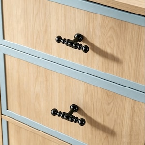 May include: Close-up of a wooden cabinet with three drawers. Each drawer has a light wood grain finish, framed by a light blue border. Black, decorative T-shaped drawer pulls are attached to each drawer, adding a touch of elegance.