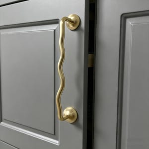 May include: A brass-coloured cabinet handle with a distinctive wavy design. The handle is mounted on a grey cabinet door with a recessed panel. The hardware features circular mounts at each end and a curved, undulating grip.