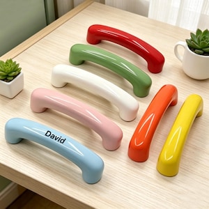 Peut inclure: Sept poignées d'armoire colorées en rouge, vert, blanc, rose, orange, jaune et bleu clair. La poignée bleu clair porte le nom "David". Les poignées sont disposées sur une surface en bois de couleur claire.