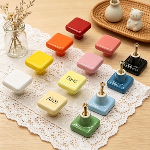 May include: A collection of colorful square ceramic cabinet knobs in various shades, including red, orange, yellow, pink, blue, and green. Some knobs are labeled with the names "David" and "Alice". Each knob has a metal screw for installation.
