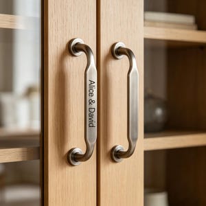 May include: Close-up of two cabinet doors with brushed metal handles. The handles are curved and feature the engraved text "Alice & David". The doors are made of light-colored wood.