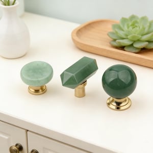 May include: Three decorative cabinet knobs with green stone tops and brass bases. The knobs have different shapes: a round, a faceted rectangular, and a spherical design. The knobs are displayed on a white surface.