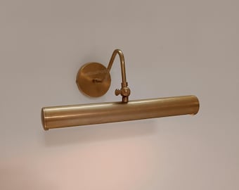 May include: A brass-toned picture light fixture. The fixture has a circular wall mount, a curved arm, and a cylindrical light housing. The light is mounted on a white wall. The fixture is designed to illuminate artwork.