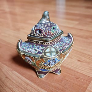 Peut inclure: Un brûleur d'encens décoratif en céramique ornée avec un couvercle. Le brûleur présente une base marron avec des bordures dorées et des motifs floraux et géométriques colorés. Le couvercle est surmonté d'un fleuron doré.