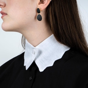 May include: A white, scalloped-edge collar is buttoned to a black garment. The model is wearing black oval earrings with gold-colored trim. The earrings are hanging from a gold-colored hoop earring.