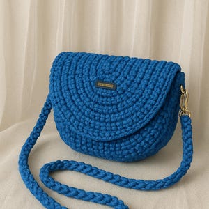 May include: A vibrant blue crochet crossbody bag with a rounded flap and a matching braided strap. The bag features a gold-colored clasp and a small rectangular tag with text. The bag is made of thick, textured yarn.