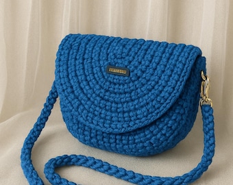 May include: A bright blue crochet crossbody bag with a rounded flap and a matching braided strap. The bag features a gold-coloured clasp and a small rectangular tag with text. The bag is made of thick, textured yarn.
