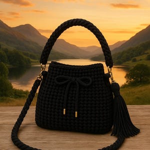 Peut inclure: Un sac à main noir en crochet avec une poignée tressée et une longue bandoulière réglable. Le sac est doté d'une fermeture à cordon avec un nœud et d'un pompon décoratif. Des détails dorés ornent le sac. Le sac est posé sur un fond panoramique.