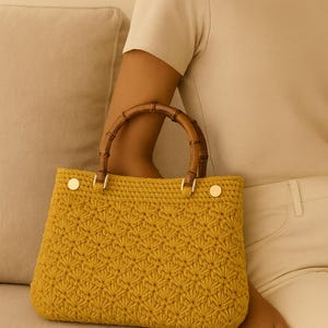 May include: Mustard yellow crochet handbag with a bamboo handle. The bag features a textured pattern and gold-colored hardware. The handle is a curved, natural bamboo. The bag is a structured tote style.
