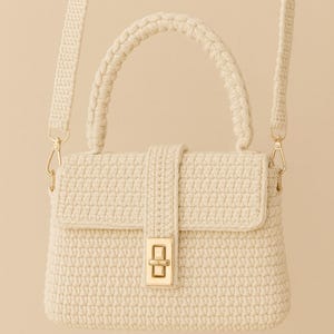 May include: Cream-colored crochet handbag with a flap closure and a gold-tone clasp. The bag features a braided top handle and a detachable shoulder strap. The crochet pattern is visible throughout the bag's design.