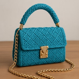 May include: A teal crochet handbag with a gold chain strap and handle. The bag features a diamond pattern and a gold clasp. The handle is a thick, braided design. The bag is sitting on a wooden surface.