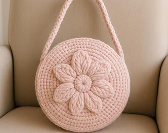 May include: A round, light pink crochet bag with a large flower design on the front. The bag has a braided handle and is sitting on a beige chair. The flower is the same color as the bag.