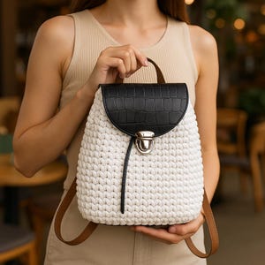 Może przedstawiać: Biały plecak z szydełka z czarną, teksturowaną klapką i brązowym uchwytem. Plecak ma brązowe paski i srebrne zapięcie. Torba jest trzymana przez osobę.