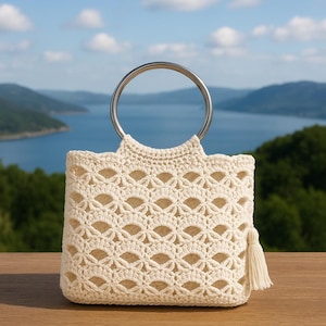 Könnte beinhalten: Cremefarbene Häkeltasche mit silberfarbenem Metallgriff. Die Tasche hat ein dekoratives Lochmuster und eine Quaste. Der Hintergrund zeigt eine malerische Aussicht auf einen See und Berge unter blauem Himmel.