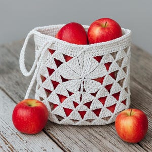 May include: A white, crocheted basket filled with red apples. The basket has a geometric design with triangular cutouts, and a handle. Three red apples are on the wooden surface next to the basket.