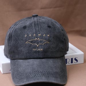 Puede incluir: Gorra de béisbol gris oscuro con el texto "DAD MAN" sobre un símbolo de murciélago dorado y "EST 2026" debajo. La gorra tiene una visera curva y un diseño cosido.