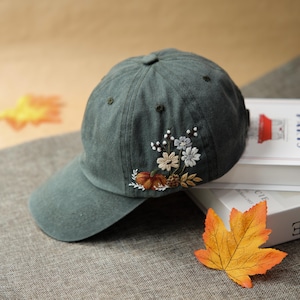 以下が含まれることがあります： 刺繍の花柄が施された濃い緑色の野球帽。刺繍には、カボチャ、松ぼっくり、オレンジ、白、青の色合いの花が描かれています。帽子にはカーブしたつばと、上部にボタンが付いています。