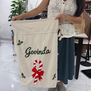 May include: A cream-colored fabric drawstring sack featuring the name Govinda in dark green script, a red and white striped candy cane with a large red bow, and small green holly leaf accents with red berries on the front.