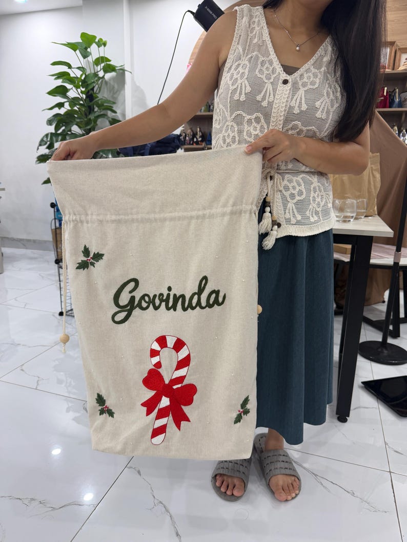 May include: A cream-colored fabric drawstring sack featuring the name Govinda in dark green script, a red and white striped candy cane with a large red bow, and small green holly leaf accents with red berries on the front.