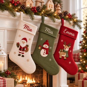 Puede incluir: Tres calcetines navideños en crema, verde oliva y rojo, cada uno con un lazo para colgar. El calcetín crema muestra a Santa Claus, el verde a un muñeco de nieve y el rojo a un reno. Cada calcetín está bordado con un nombre.