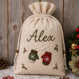 May include: A beige linen drawstring gift bag featuring the embroidered name Alex in dark green script. The bag is decorated with two small mittens, one green and one red, connected by a white string, along with embroidered holly sprigs.