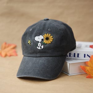 May include: A charcoal gray baseball cap featuring an embroidered image of Snoopy holding a sunflower, with Woodstock nearby. The cap has a curved brim and a button on top. The design is on the front of the cap.