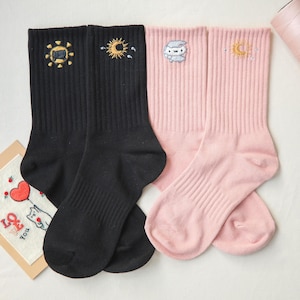 Könnte beinhalten: Zwei Paar Socken, eine schwarze und eine rosa. Die schwarzen Socken haben gestickte Sonnenmotive, während die rosa Socken eine gestickte Eule und Sonne haben. Die Socken sind aus weichem Stoff.