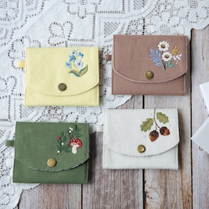Könnte beinhalten: Vier kleine, quadratische Stoffbeutel in Pastellfarben: Gelb, Braun, Grün und Weiß. Jeder Beutel hat ein gesticktes Blumen- oder Naturmotiv, darunter Vergissmeinnicht, Gänseblümchen, Eicheln und ein Pilz. Jeder hat einen Knopfverschluss.