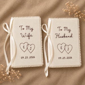 Può includere: Due libri in tessuto color avorio con bordi sfrangiati e nastri color crema. Il libro a sinistra è ricamato con "To My Wife" e due cuori contenenti le lettere "E" e "J" sopra la data "09.25.2026". Il libro a destra riporta "To My Husband".