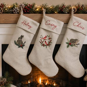 May include: Three beige fabric Christmas stockings with embroidered cuffs. The first features a rabbit, the second a wreath with two red birds, and the third a hedgehog. Each cuff is personalized with the names Haley, Casey, and Chris in red.