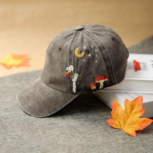 May include: A brown baseball cap with embroidered whimsical designs. The cap features a crescent moon, stars, mushrooms, and flowers. The brim is curved, and the cap has a button on top. The cap is sitting on a grey surface.