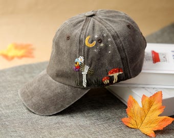 Op de afbeelding: Een bruine baseballcap met geborduurde grillige ontwerpen. De cap heeft een halve maan, sterren, paddenstoelen en bloemen. De klep is gebogen en de cap heeft een knoop aan de bovenkant. De cap ligt op een grijze ondergrond.