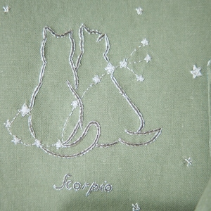 Peut inclure: Tissu vert sauge avec un motif brod&eacute; de deux silhouettes de chats et une constellation, avec le mot "Scorpio" en dessous. Le motif est cousu avec du fil blanc. De petits ornements en forme d'&eacute;toiles sont dispers&eacute;s sur le tissu.