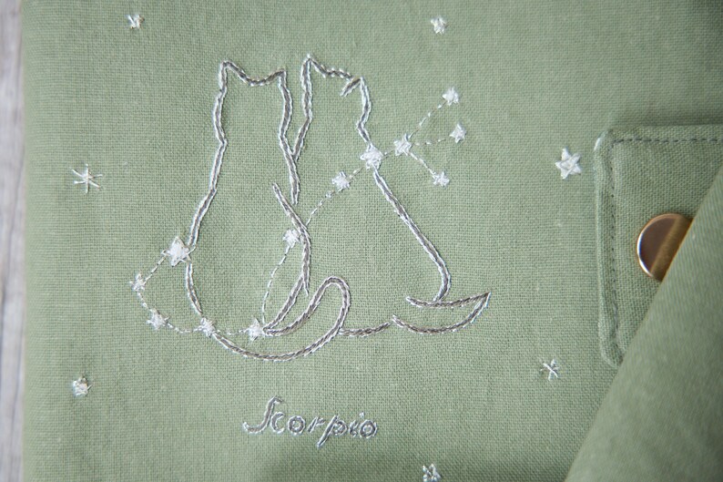 Peut inclure: Tissu vert sauge avec un motif brod&eacute; de deux silhouettes de chats et une constellation, avec le mot "Scorpio" en dessous. Le motif est cousu avec du fil blanc. De petits ornements en forme d'&eacute;toiles sont dispers&eacute;s sur le tissu.