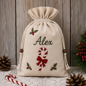 May include: A beige drawstring gift bag made of fabric featuring the embroidered name Alex in green script. The bag is decorated with a red and white candy cane, a red bow, and green holly leaves with red berries, finished with white cord ties.