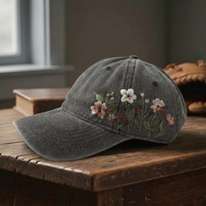 Op de afbeelding: Een donkergrijze baseballcap met geborduurd bloemmotief. De bloemen zijn in tinten roze, wit en bruin met groene bladeren. De cap heeft een gebogen klep en een knoop aan de bovenkant. Geplaatst op een houten oppervlak.