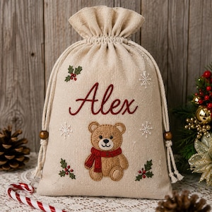 May include: A beige drawstring gift bag made of fabric featuring the embroidered name Alex in red script. The bag is decorated with a central brown teddy bear wearing a red scarf, surrounded by embroidered white snowflakes and green holly sprigs.