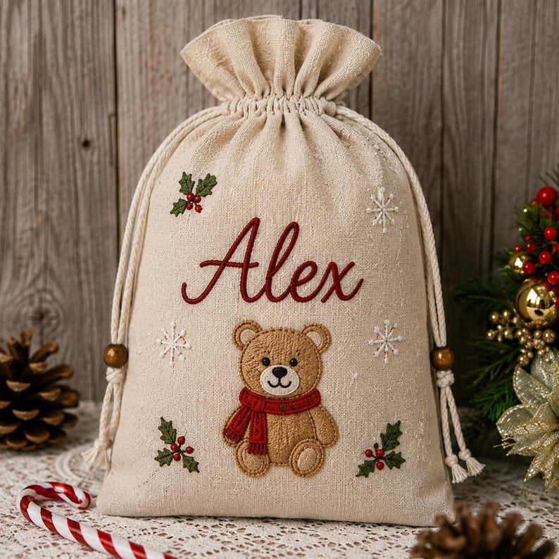 May include: A beige drawstring gift bag made of fabric featuring the embroidered name Alex in red script. The bag is decorated with a central brown teddy bear wearing a red scarf, surrounded by embroidered white snowflakes and green holly sprigs.